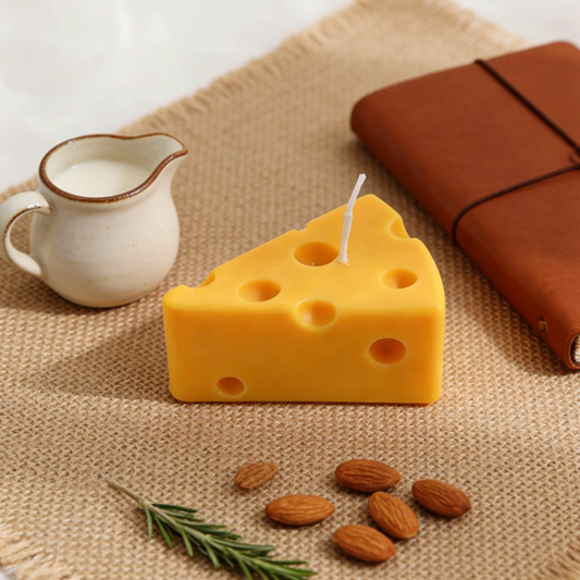 Realistic Swiss cheese wedge-shaped candle in bright yellow, styled in a cozy table setting with a ceramic milk jug, brown leather notebook, almonds, and rosemary on a burlap mat