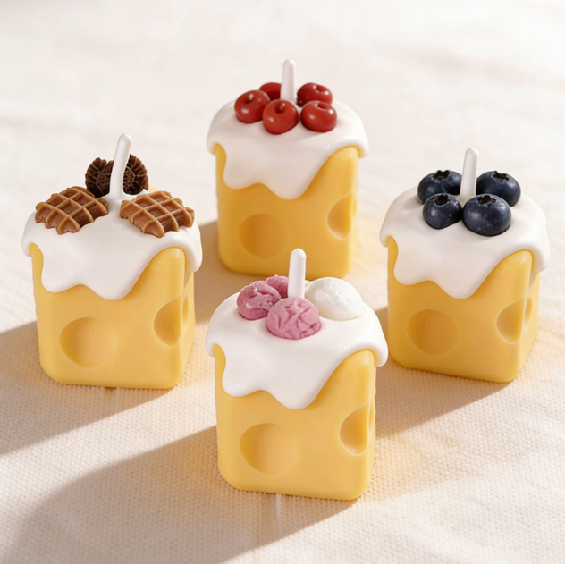 Assortment of realistic cheese-shaped candles with white cream wax drizzle, topped with decorative waffle, cookie, cherry, blueberry, and ice cream wax sculptures, displayed on a neutral background