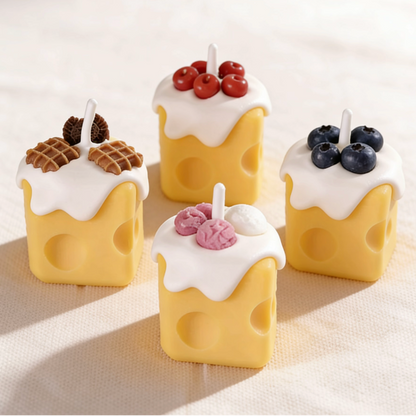 Assortment of realistic cheese-shaped candles with white cream wax drizzle, topped with decorative waffle, cookie, cherry, blueberry, and ice cream wax sculptures, displayed on a neutral background