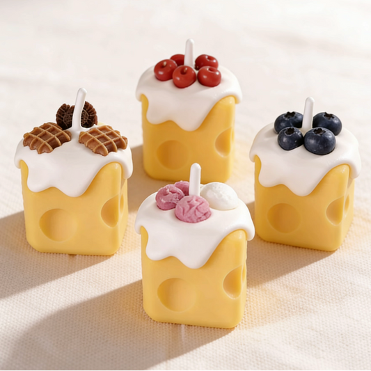 Assortment of realistic cheese-shaped candles with white cream wax drizzle, topped with decorative waffle, cookie, cherry, blueberry, and ice cream wax sculptures, displayed on a neutral background