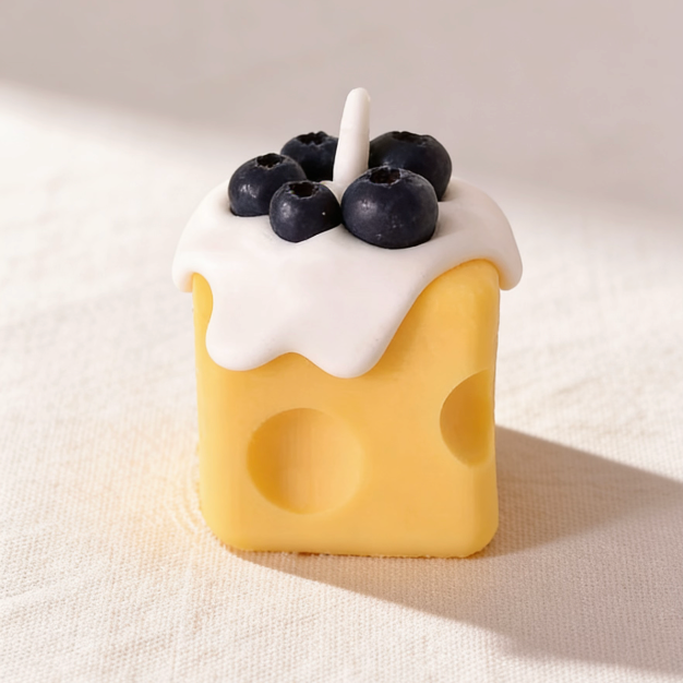 Realistic cheese-shaped candle with a yellow Swiss cheese design, topped with white cream wax drizzle and decorative blueberry wax sculptures, featuring a cotton wick, displayed on a neutral background