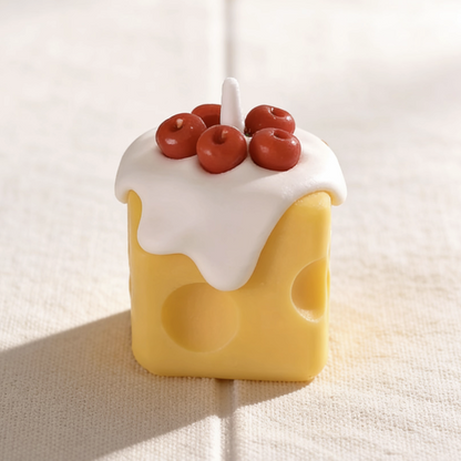 Realistic cheese-shaped candle with a yellow Swiss cheese design, topped with white cream wax drizzle and decorative cherry wax sculptures, featuring a cotton wick, displayed on a neutral background