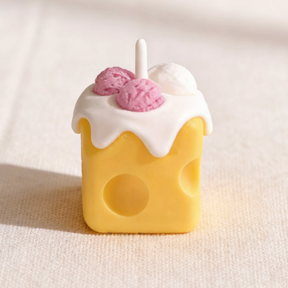 Realistic cheese-shaped candle with a yellow Swiss cheese design, topped with white cream wax drizzle and decorative pink and white ice cream scoop wax sculptures, featuring a cotton wick, displayed on a neutral background