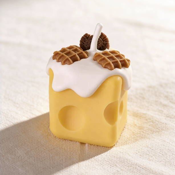 Realistic cheese-shaped candle with a yellow Swiss cheese design, topped with white cream wax drizzle and decorative waffle and cookie wax sculptures, featuring a cotton wick, displayed on a neutral background