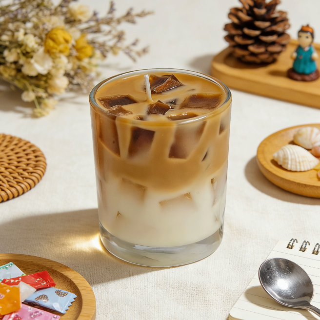 Layered iced coffee candle in a clear glass tumbler, with a white milk base, brown coffee layer, and realistic ice cube wax sculptures, styled in a cozy table setting with dried flowers, pinecone, seashells, and small decor items