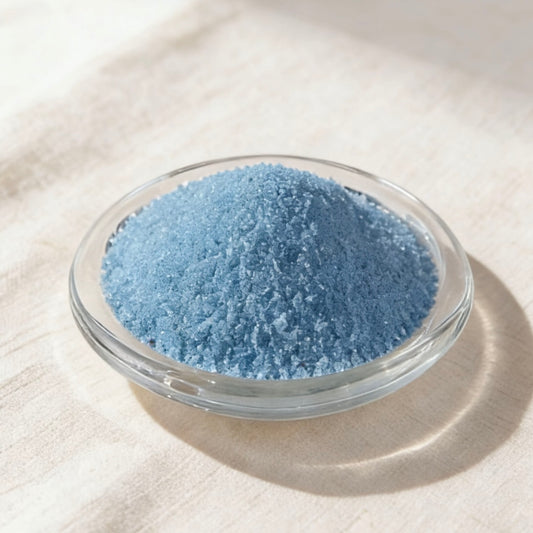 英文：Light blue candle wax granules in a clear glass bowl on a white linen background
