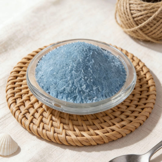 Light blue candle wax powder in a glass bowl on a rattan placemat, with a white seashell accent
