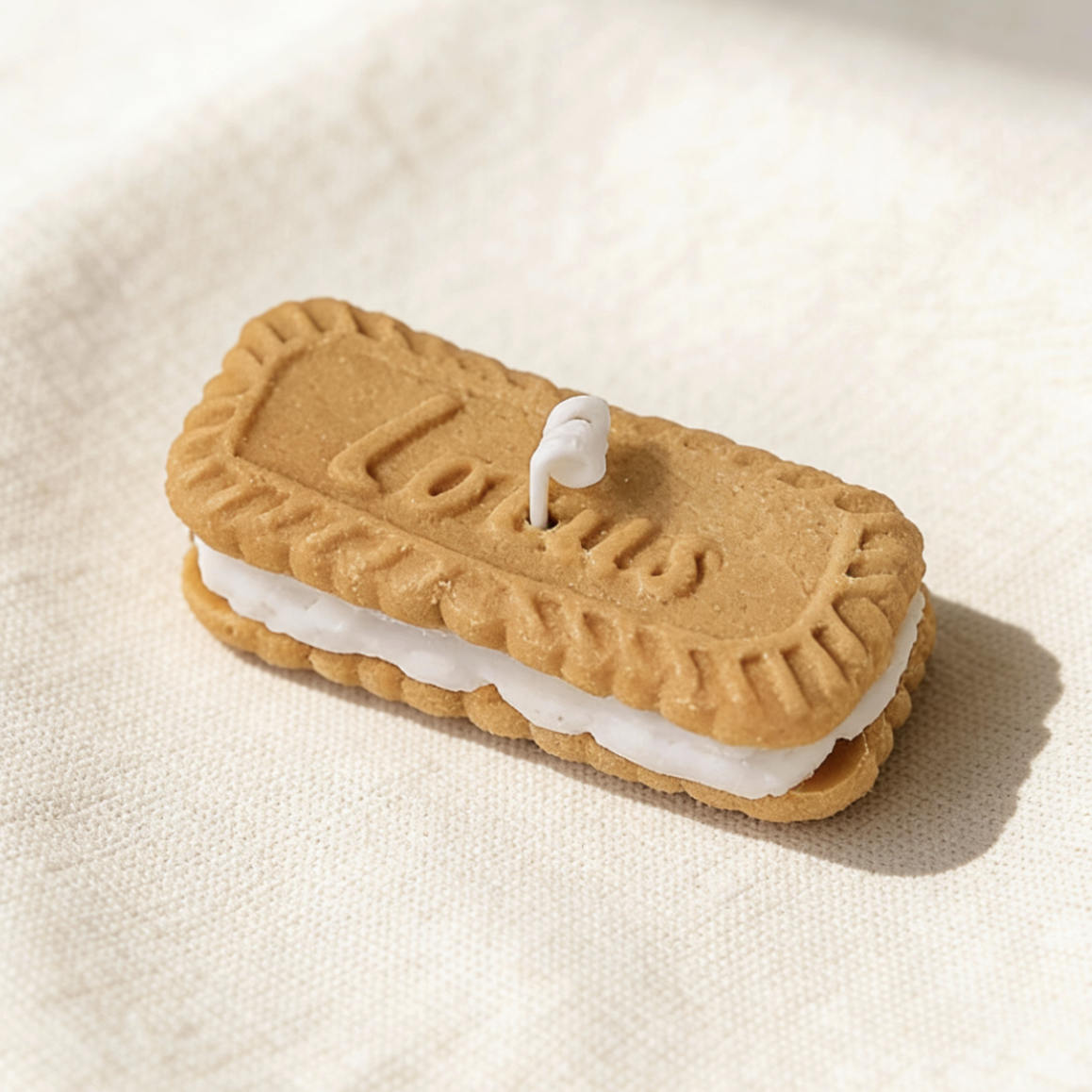 Realistic sandwich cookie-shaped candle, styled after Lotus Biscoff biscuits, with golden-brown embossed cookie layers, white cream filling, and an embossed "Lotus" logo, featuring a cotton wick, displayed on a neutral background