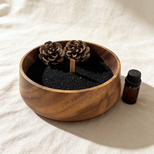 Decorative sand candle in a round wooden bowl, filled with black sand, topped with two pinecones, and accompanied by an amber essential oil bottle on a soft white linen background