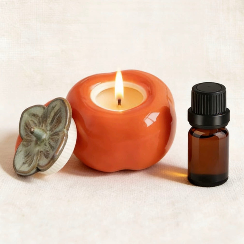 Burning candle in a glossy orange persimmon-shaped ceramic holder with a metal leaf lid, accompanied by an amber essential oil bottle on a neutral linen background