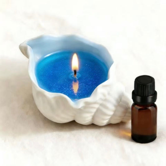 Burning blue glitter candle in a white seashell-shaped ceramic holder, with a brown essential oil bottle beside it, on a soft white background
