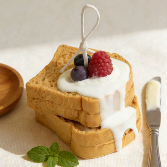 Realistic stacked toast-shaped candle with dripping white cream wax and decorative blueberry and raspberry sculptures, styled with a wooden dish, butter knife, mint leaves, and oats on a neutral background