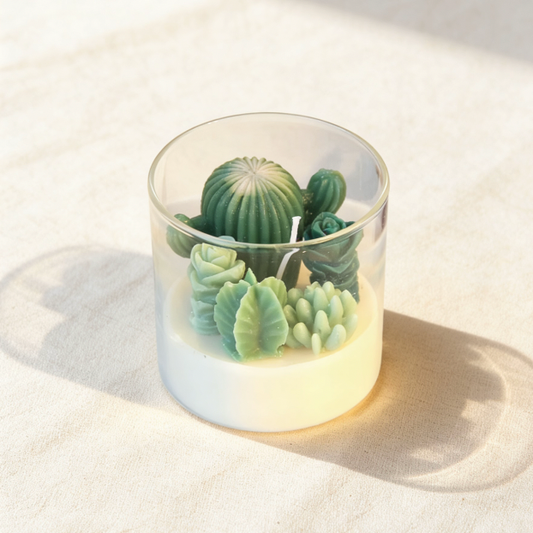 Decorative terrarium candle in a clear glass jar, featuring realistic green succulent and cactus wax sculptures embedded in a creamy white wax base
