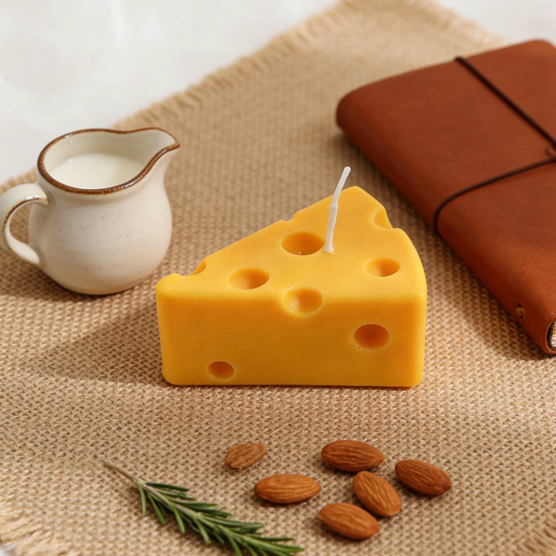 Realistic Swiss cheese wedge-shaped candle in bright yellow, styled in a cozy table setting with a ceramic milk jug, brown leather notebook, almonds, and rosemary on a burlap mat